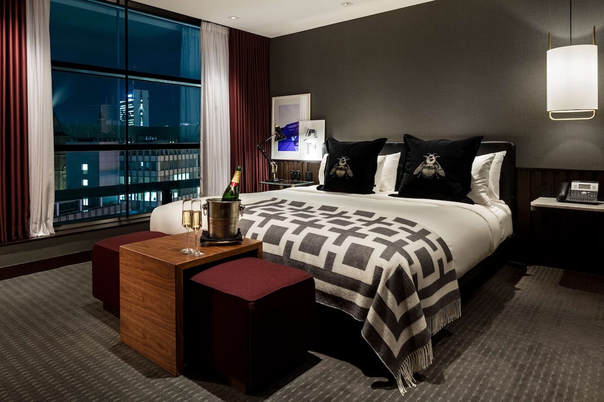 A modern hotel room with a large bed, a wooden side table with a bottle of champagne, a red ottoman, and a city view through large windows.