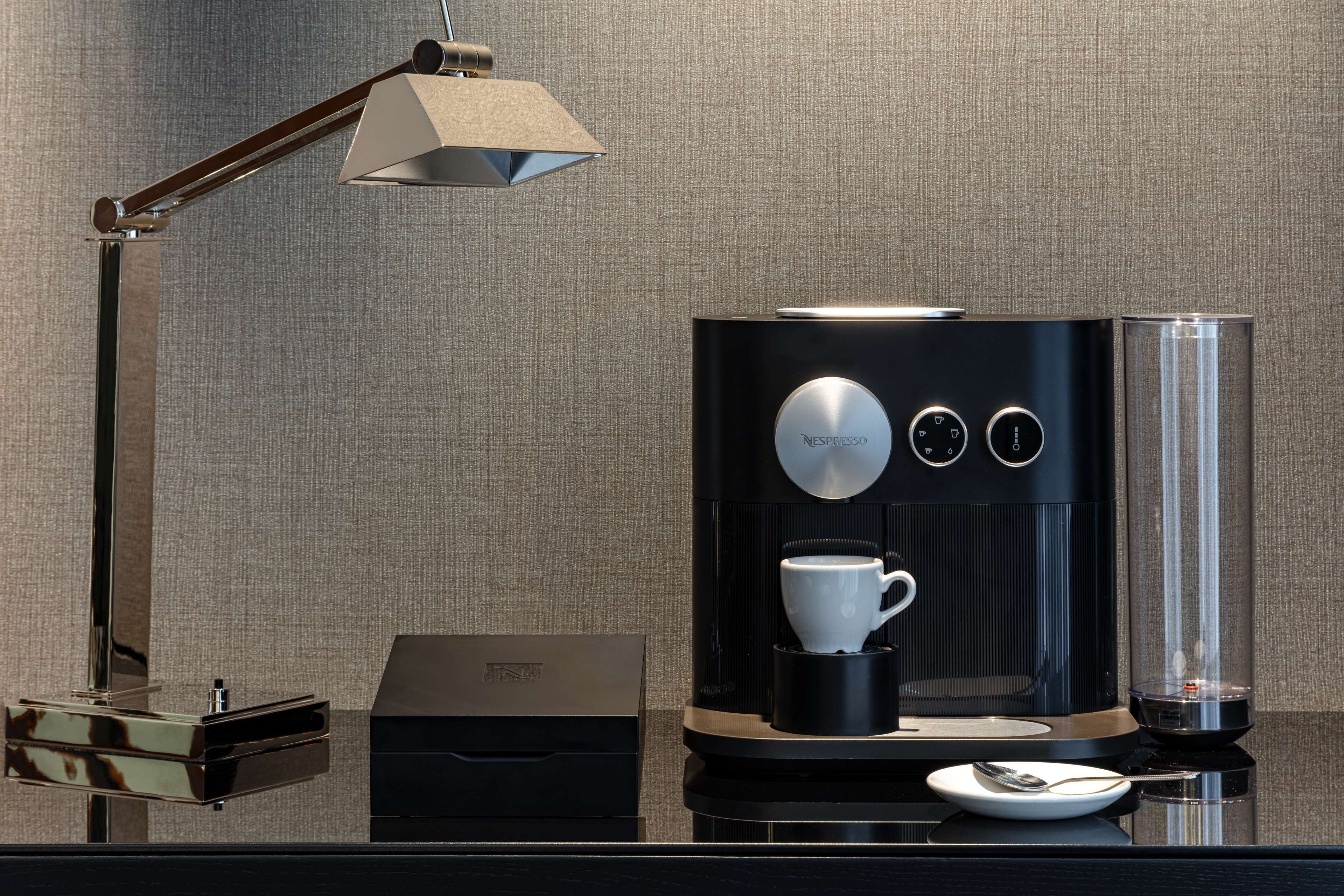 Una cafetera moderna con un porta-tazas, una cuchara y una caja, todo sobre una superficie reflectante.