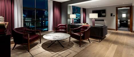 Una habitación de hotel moderna con un ventanal grande, sillones de cuero, una mesa redonda y un minibár.