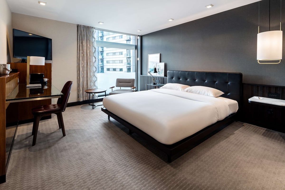 A modern hotel room with a large bed, a desk with a chair, a television, and a large window with curtains.