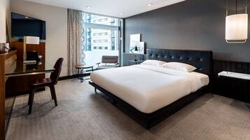 A modern hotel room with a large bed, a desk with a chair, a television, and a large window with curtains.