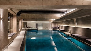 A modern indoor swimming pool with clear water, a diving board, and a lounge area with seating.