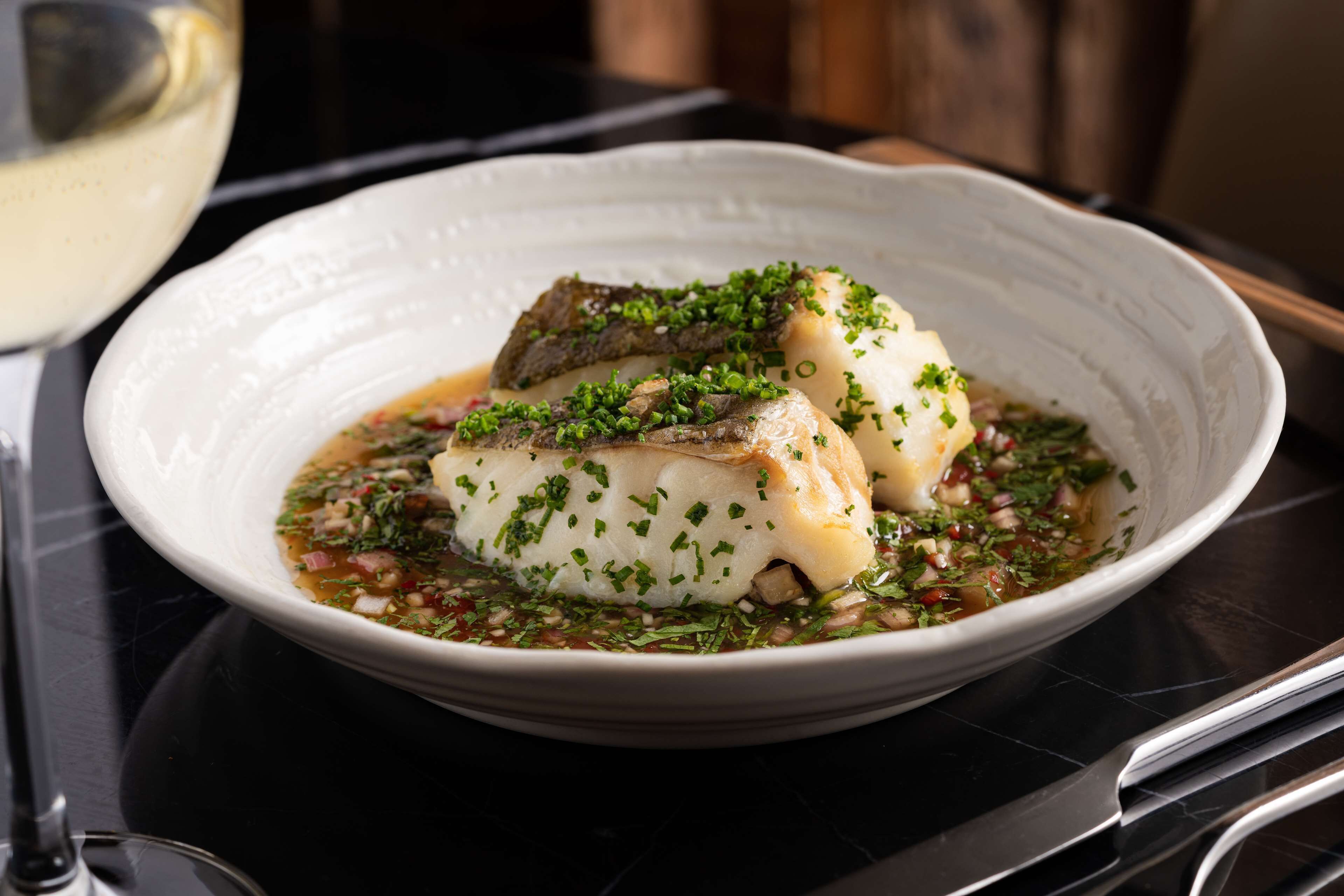 Une assiette blanche contenant deux morceaux de poisson, décorée d’herbes aromatiques, et accompagnée d’un verre de vin blanc.
