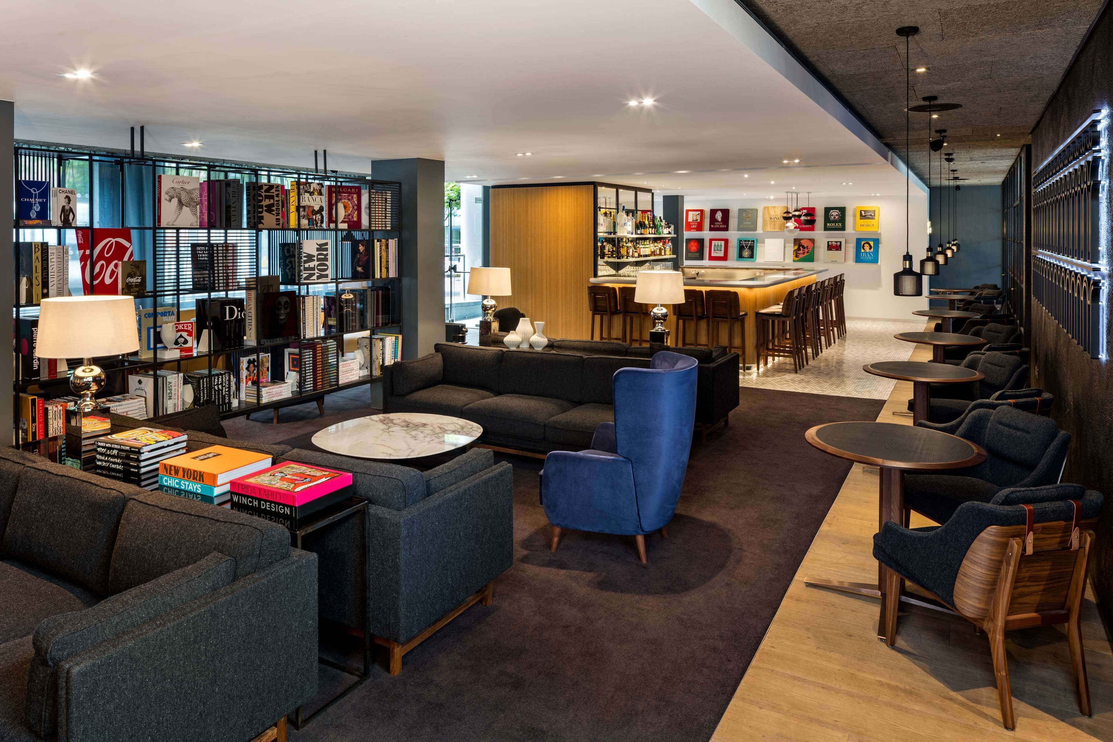 A modern lounge area with sofas, armchairs, and tables, a bar area with stools, and a bookshelf with various books and magazines.