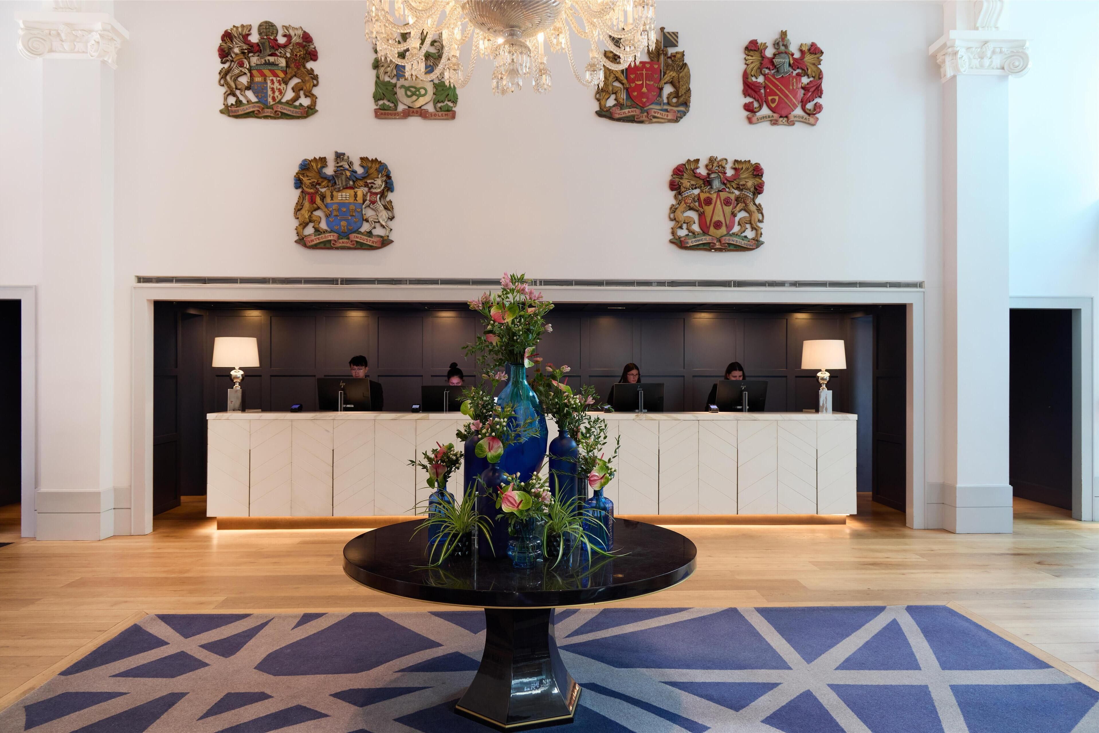 A reception area with a round table in the center, surrounded by vases with flowers. Behind the reception desk, there are coats of arms on the wall.