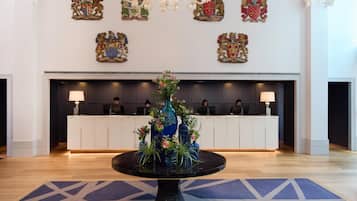A reception area with a round table in the center, surrounded by vases with flowers. Behind the reception desk, there are coats of arms on the wall.