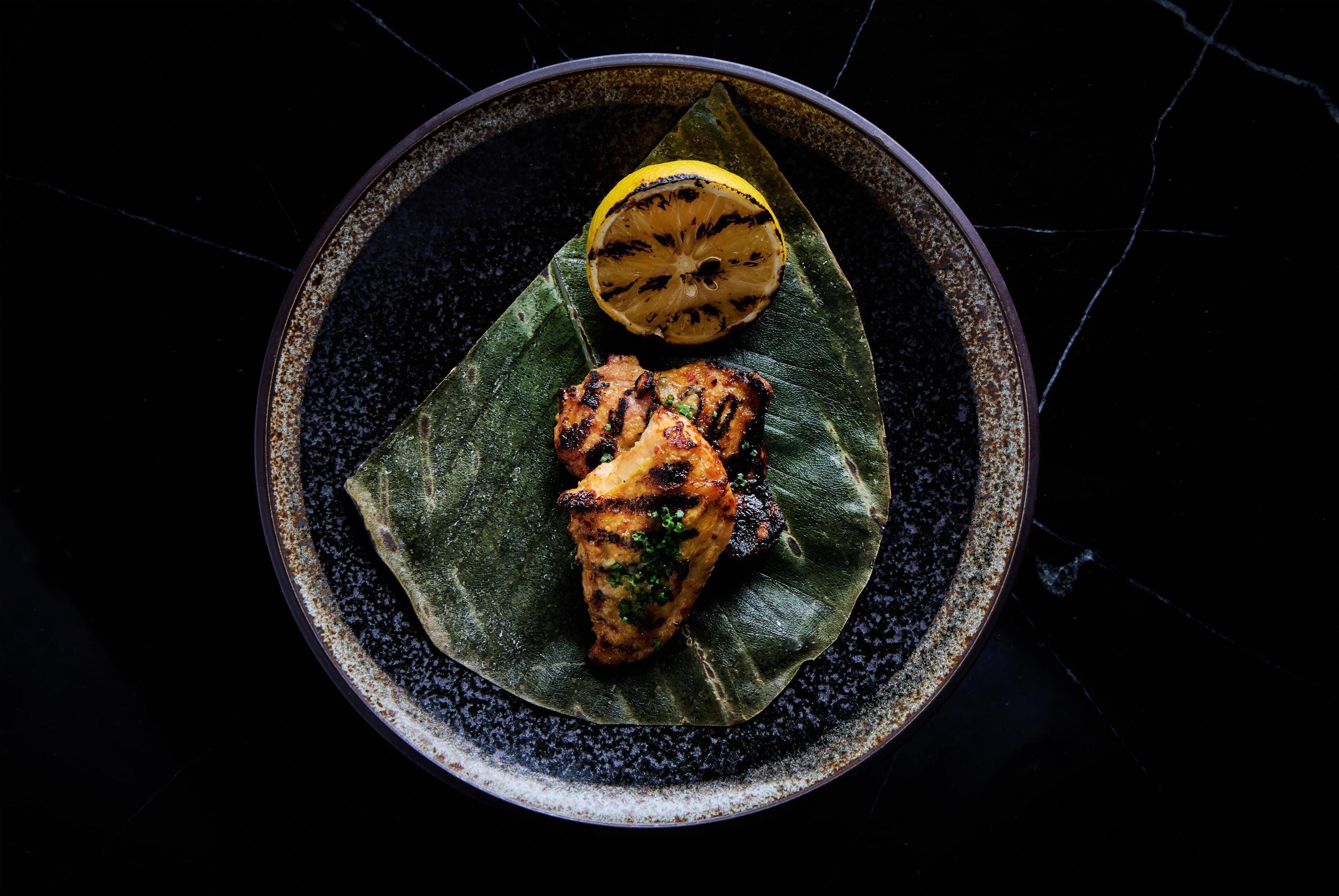 A plate with grilled chicken and a grilled lemon half on a dark speckled plate.