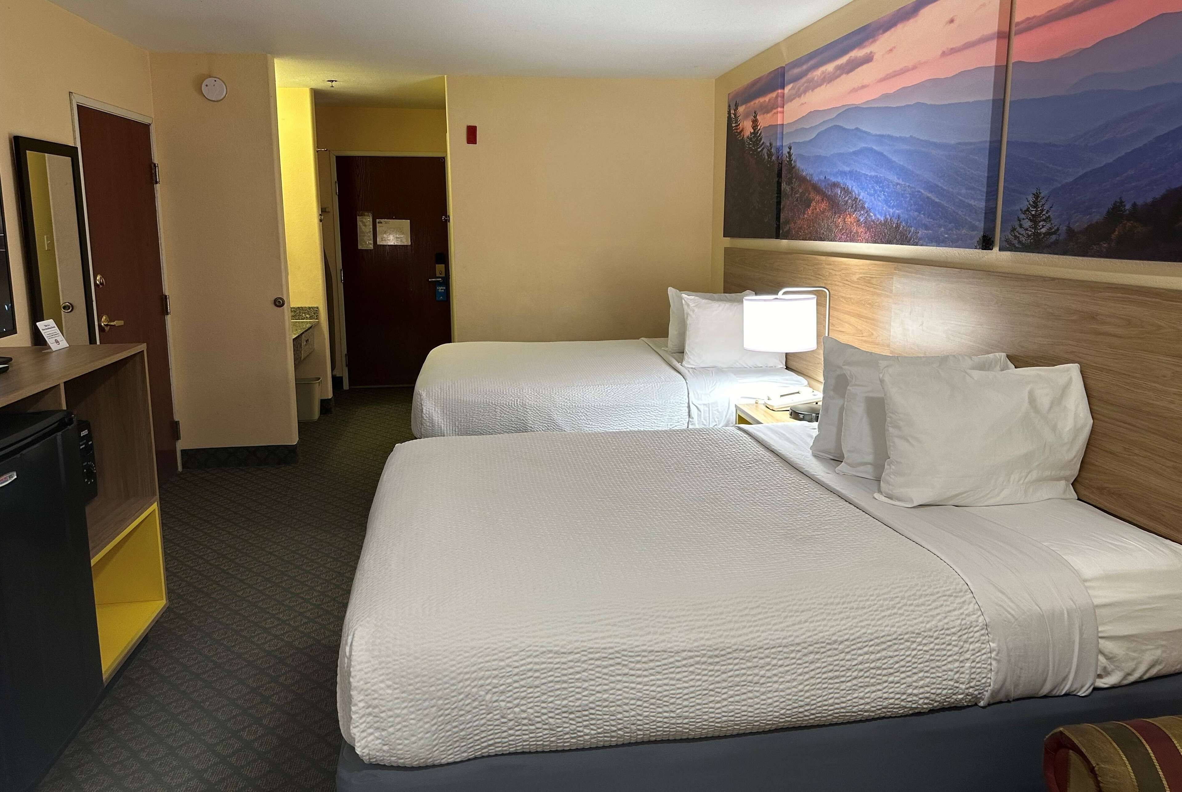 A hotel room with two beds, a desk with a lamp, a painting of a mountain landscape, and a door leading to another room.