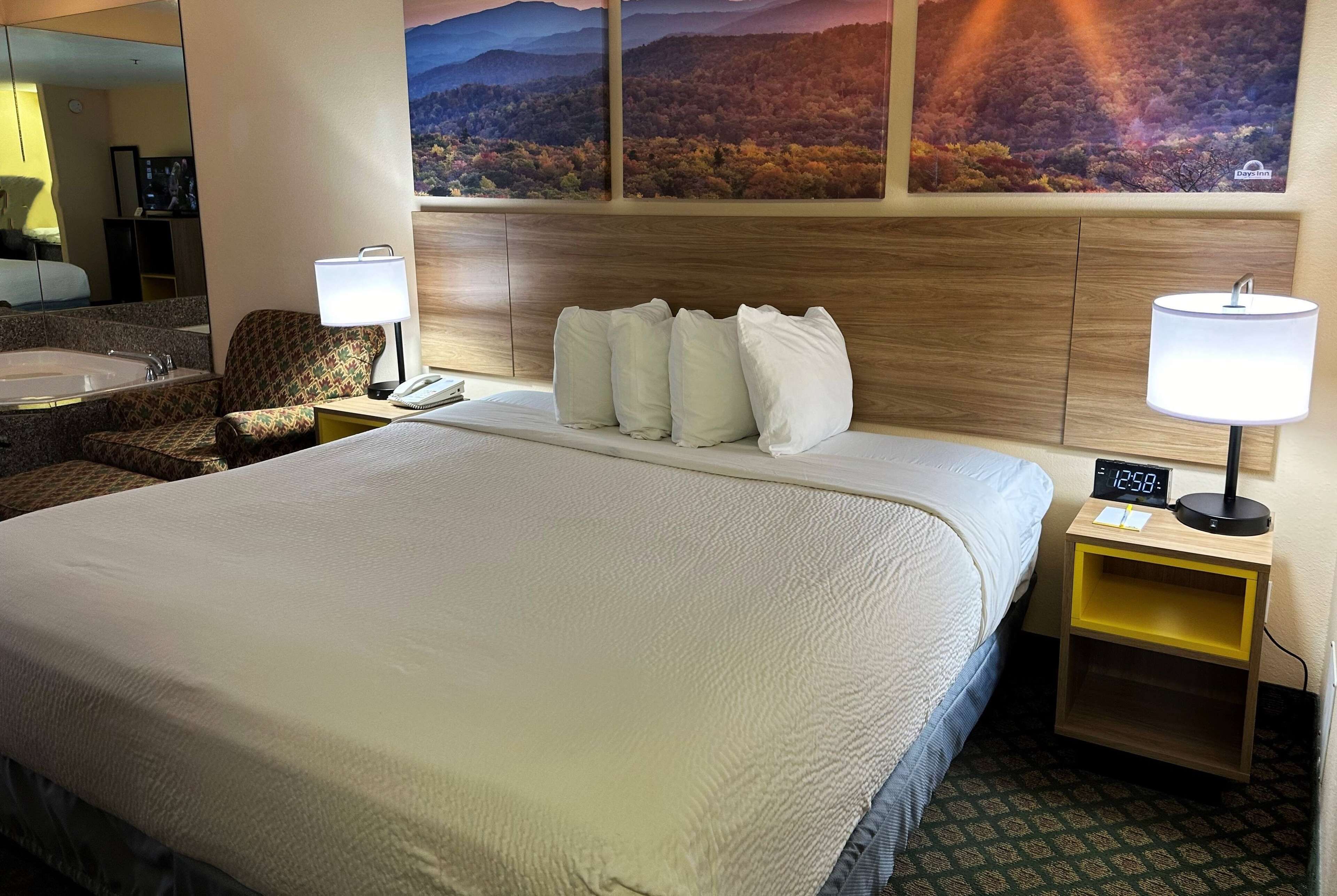 A hotel room with a large bed, two bedside tables with lamps, a chair, a mirror, and a wall with three landscape paintings.