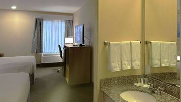 Habitación de hotel con cama, escritorio con computadora, baño con lavamanos y espejo, y ventana con cortinas.