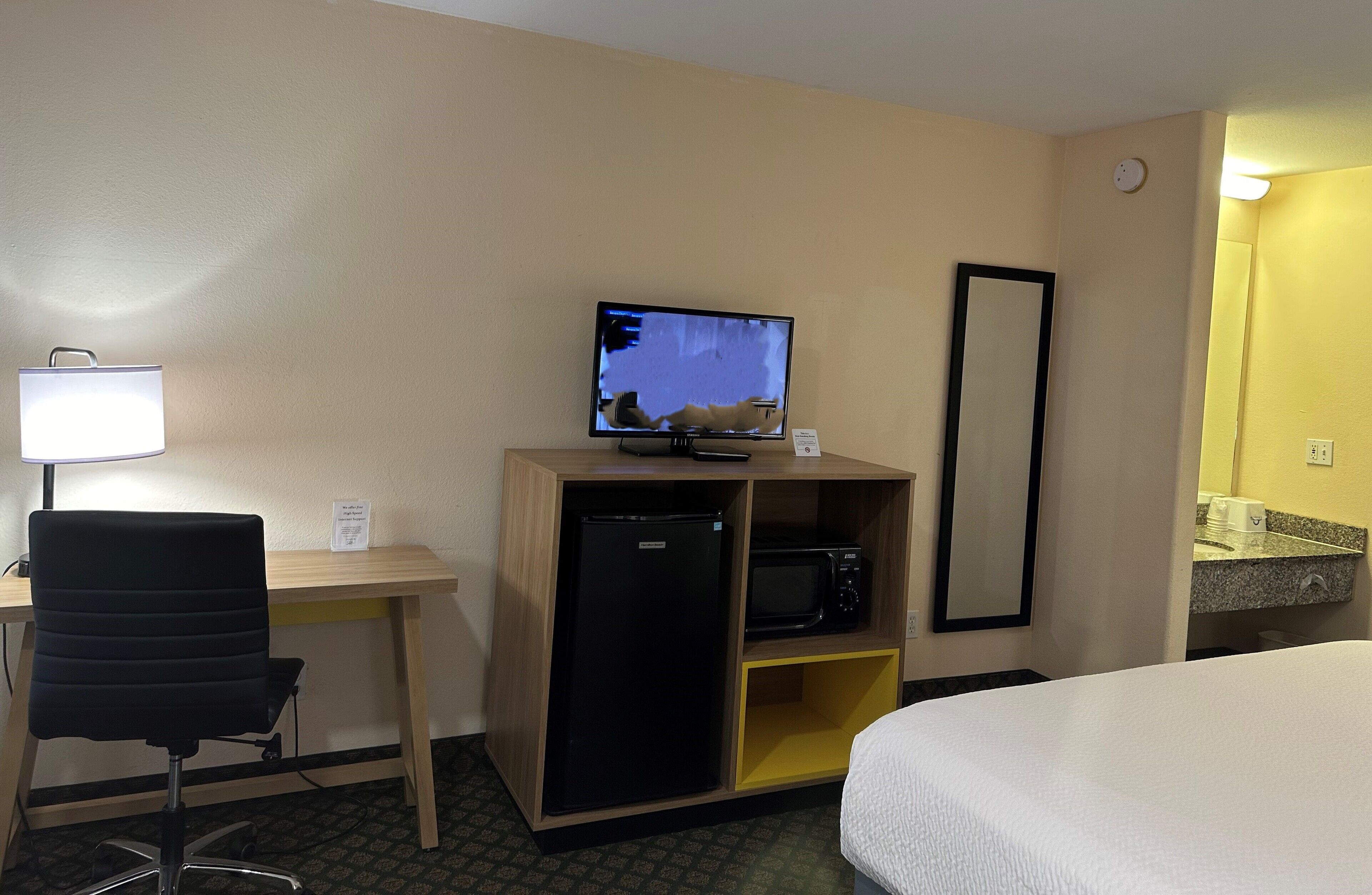 Hotelkamer met een bed, bureau met computer, koelkast, magnetron, lamp en spiegel.