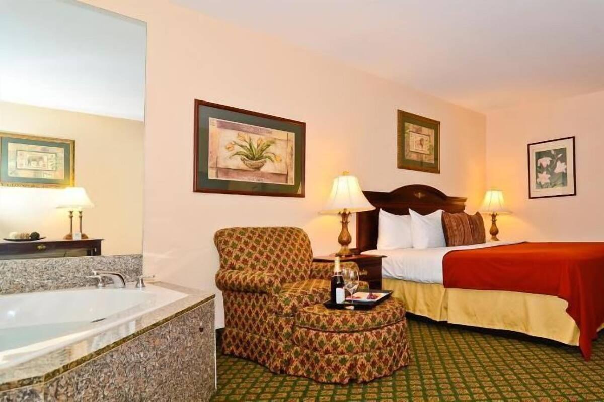 A hotel room with a bed, a chair, a desk, a lamp, a picture frame, and a bathroom with a bathtub.
