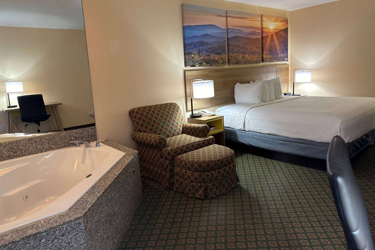 A hotel room with a large bed, a sitting area with a chair and ottoman, a desk with a computer and lamp, a large bathtub, and a painting of a mountain landscape on the wall.