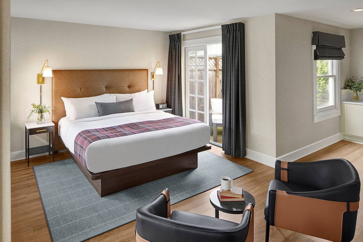 Habitació d'hotel moderna amb un llit gran, taules de nit, una cadira, una taula petita i una finestra amb cortines.