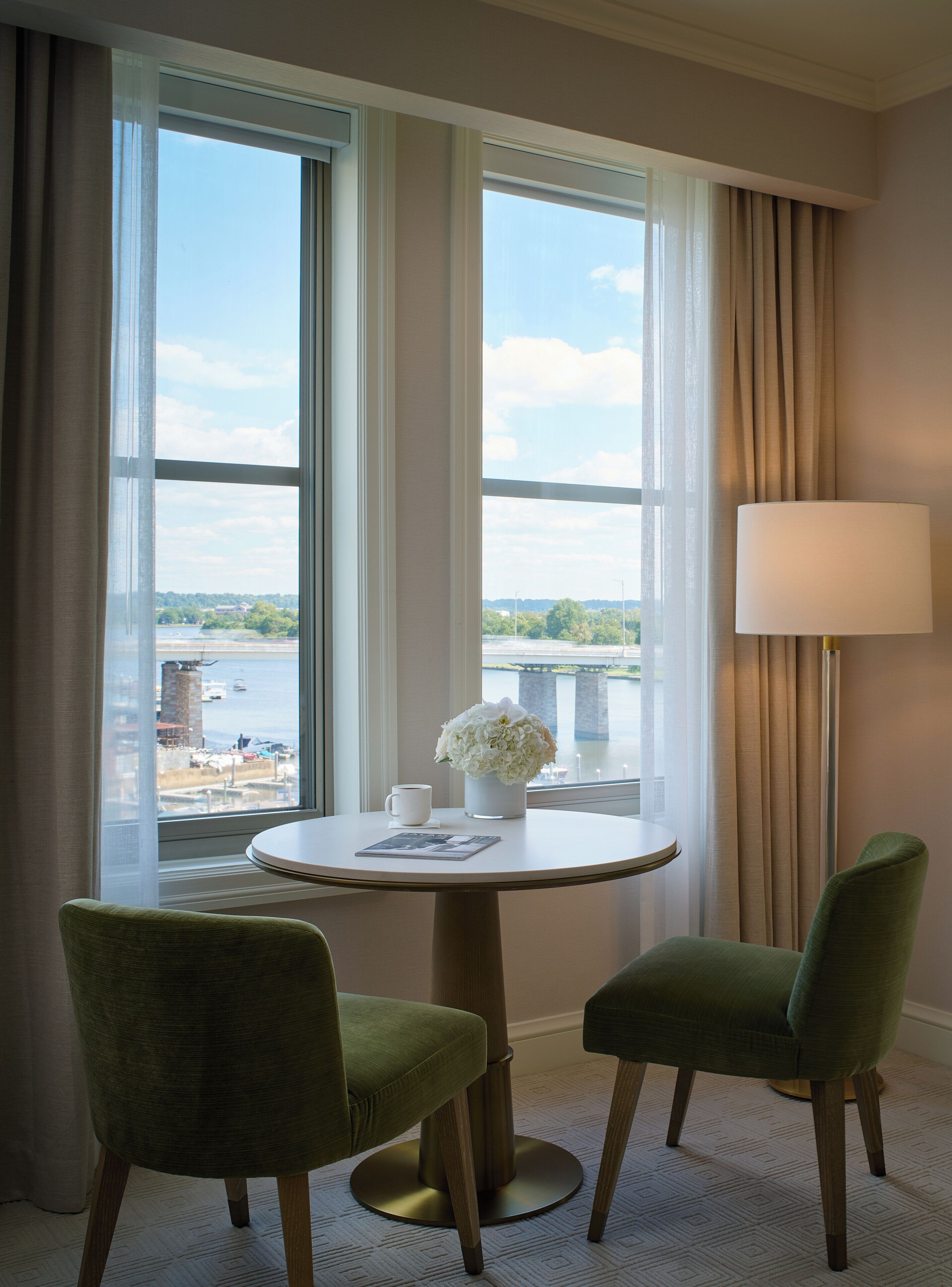 Une chambre d’hôtel avec une table ronde, deux chaises, une lampe, un vase avec des fleurs et une vue sur un pont et sur l’eau.