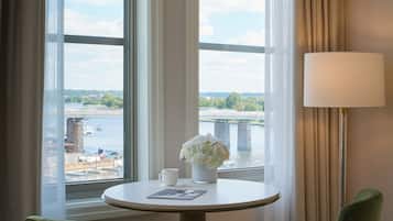 Une chambre d’hôtel avec une table ronde, deux chaises, une lampe, un vase avec des fleurs et une vue sur un pont et sur l’eau.