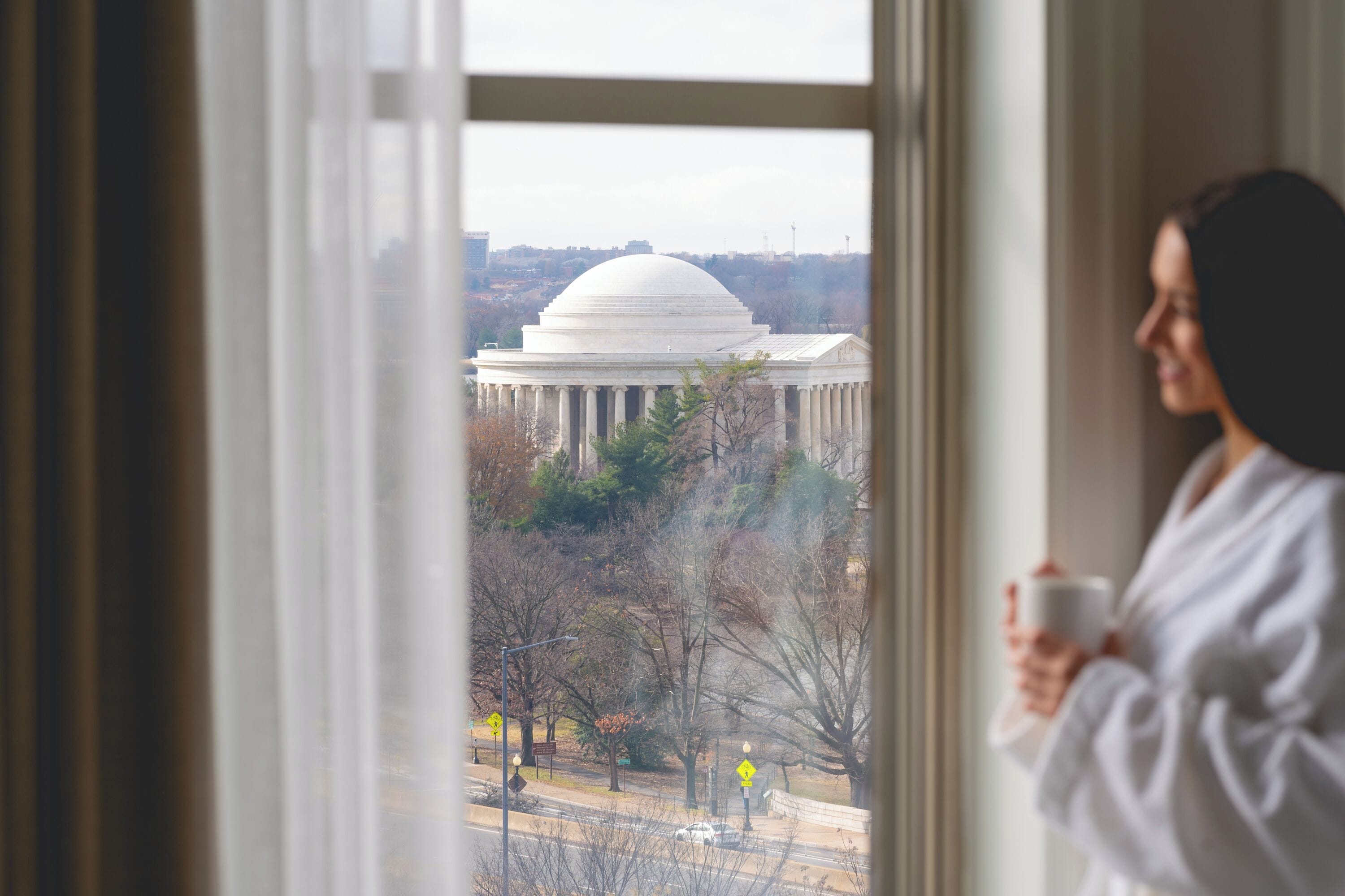 Une personne vêtue d’une robe, tenant une tasse, regarde par une fenêtre un grand bâtiment blanc orné de colonnes et surmonté d’un dôme. La scène est observée à travers une fenêtre avec des rideaux.