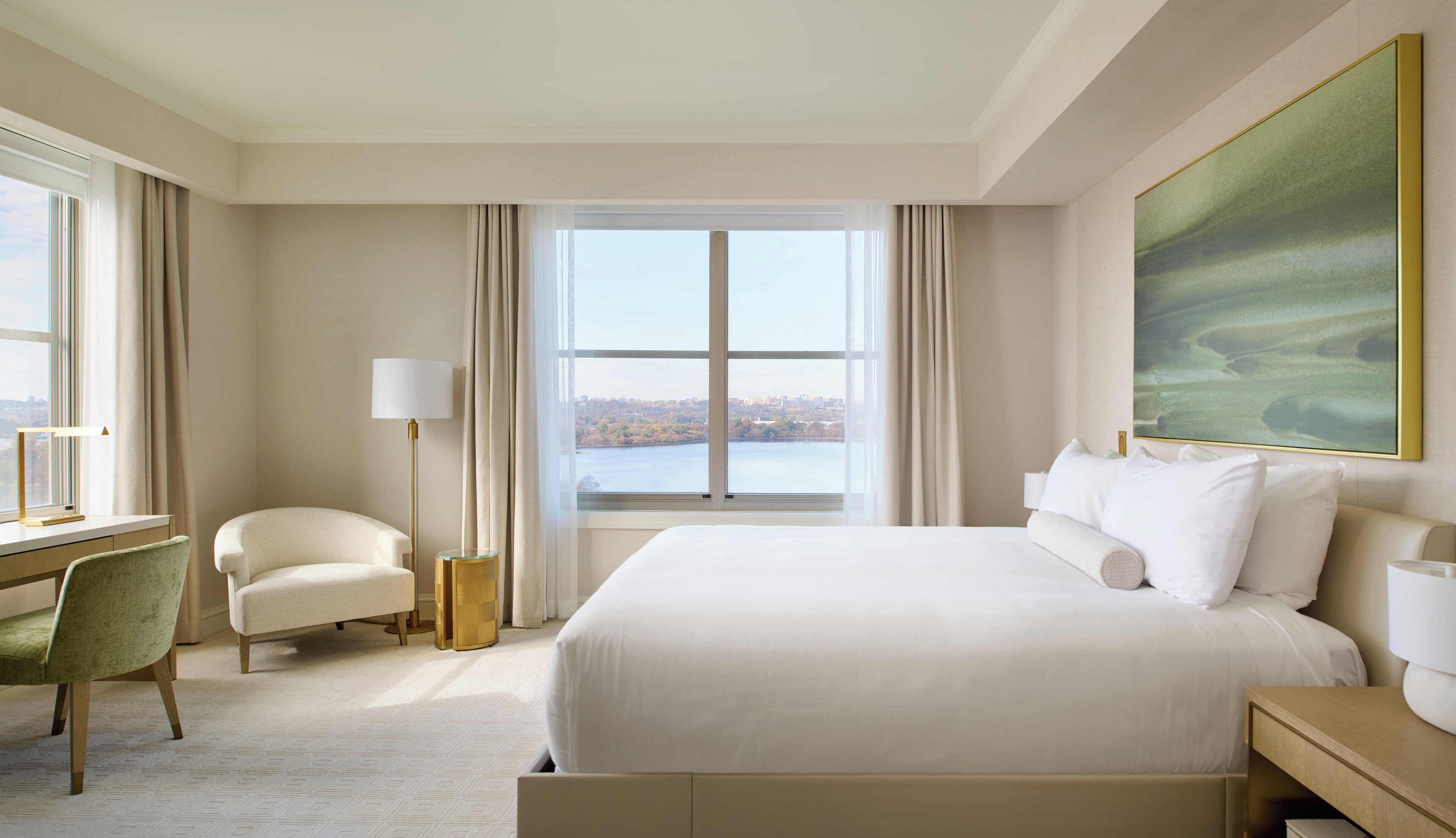 A hotel room with a large bed, a desk with a lamp, a chair, and a view of a body of water.