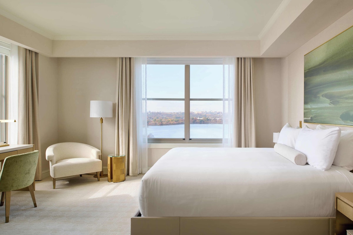 Habitación de hotel con una cama grande, un escritorio con lámpara, una silla y vistas a un cuerpo de agua.