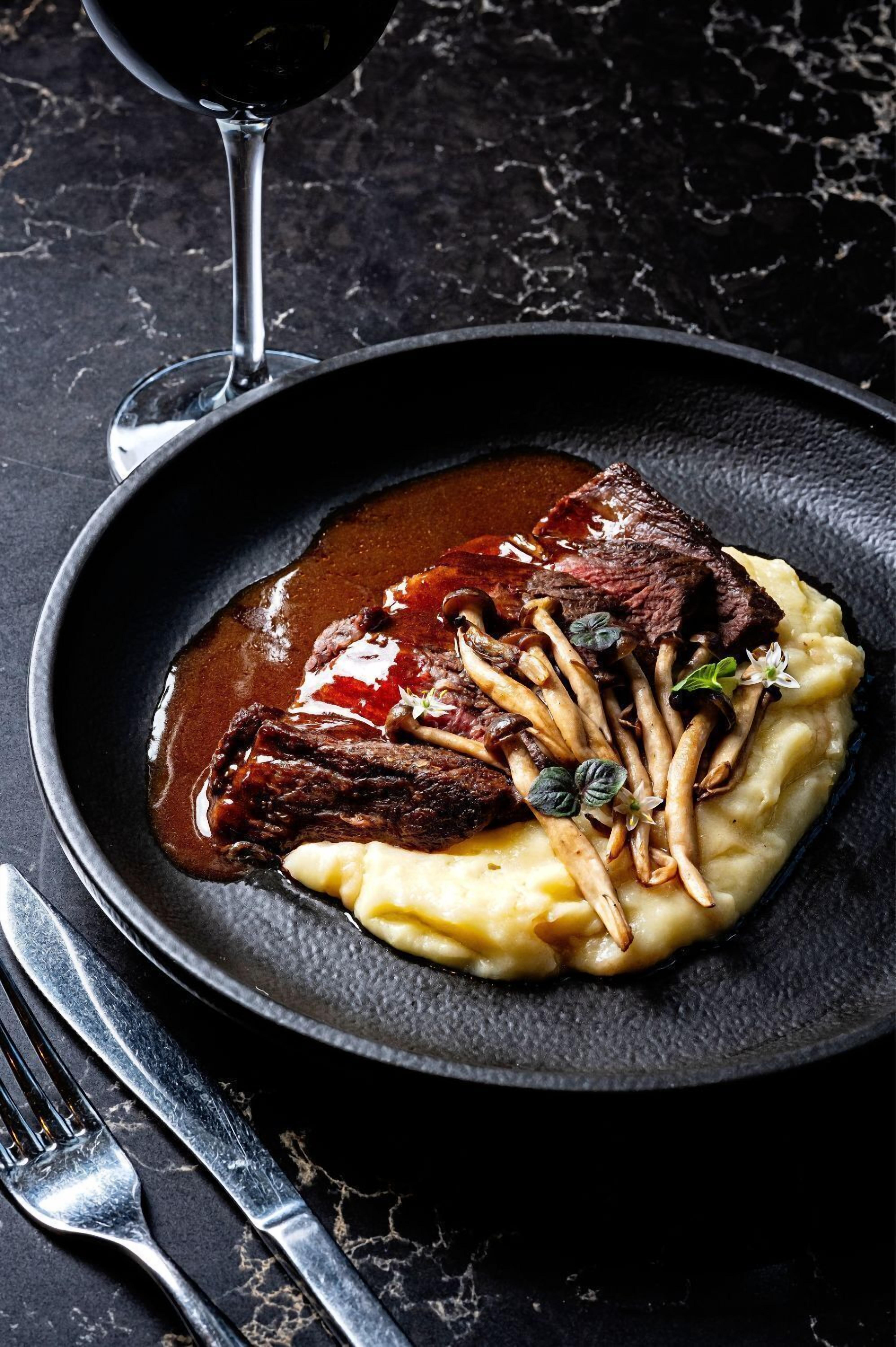 A plate of steak with mushrooms and sauce, served on a black plate with a fork and knife beside it.