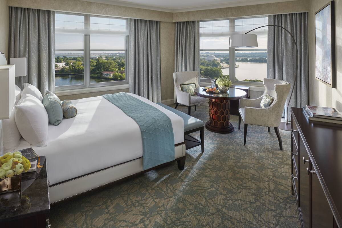 Habitación de hotel con una cama grande, dos sillas, una mesa redonda y vistas a un cuerpo de agua y un edificio.