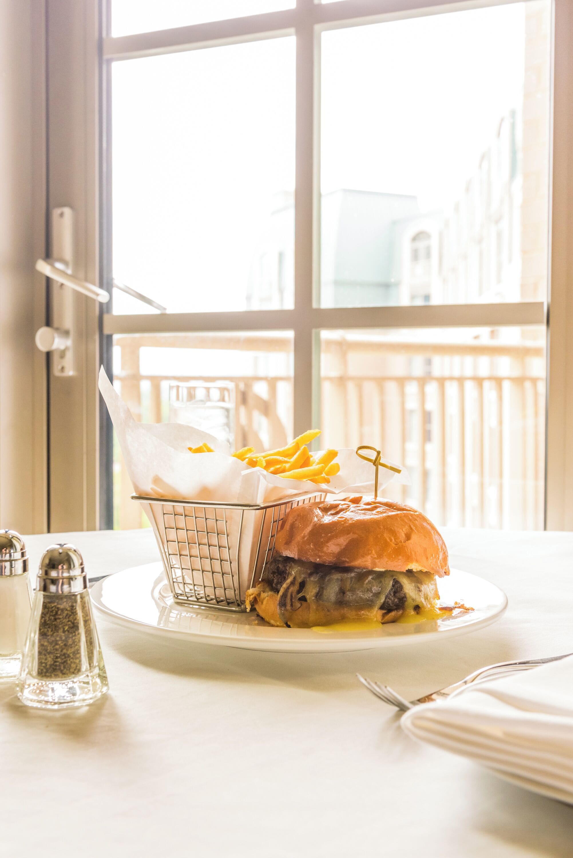 Un sándwich con papas fritas en un plato, un salero y un pimentero sobre una mesa junto a una ventana.
