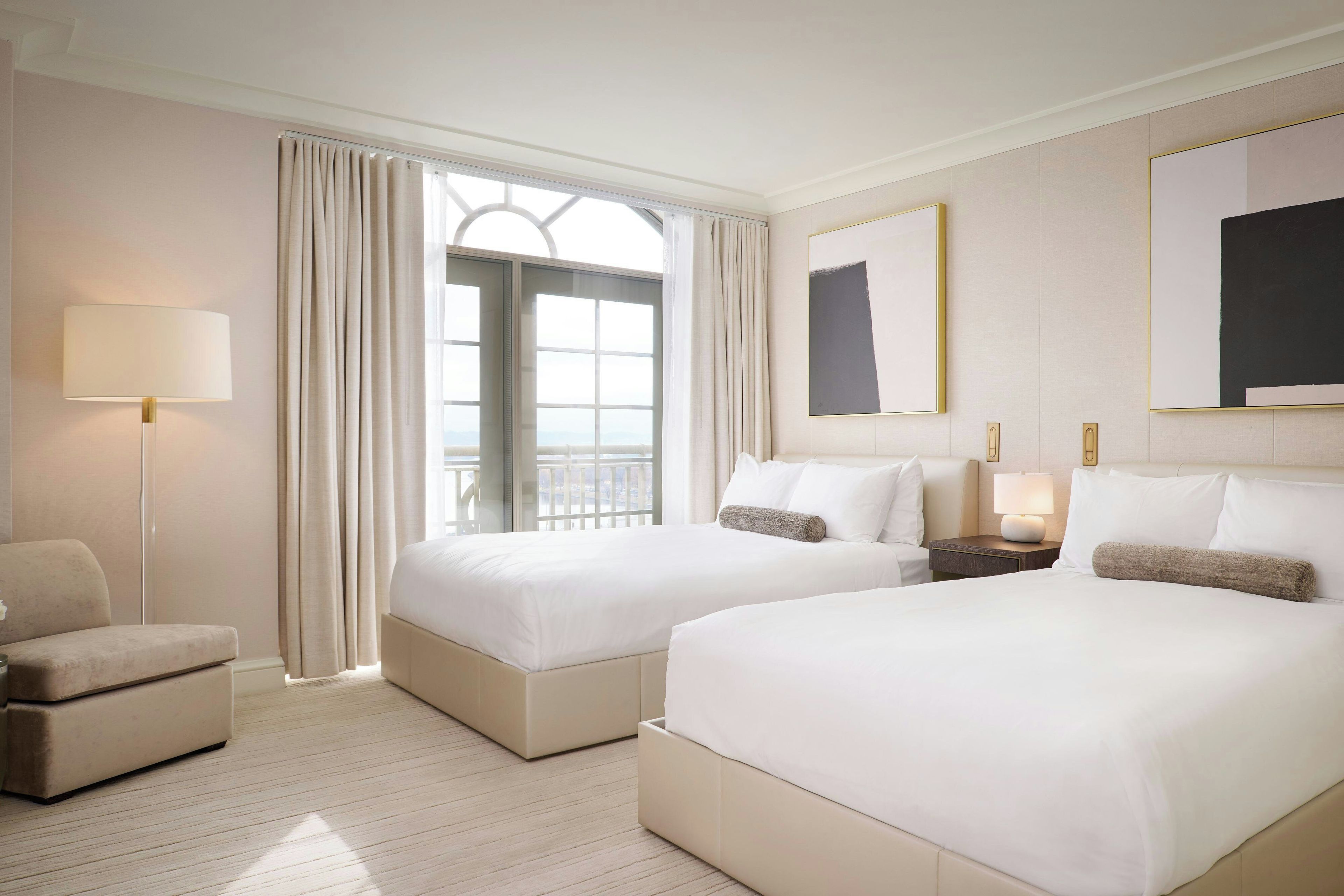 A hotel room with two beds, a chair, a nightstand with a lamp, and a large window with curtains.