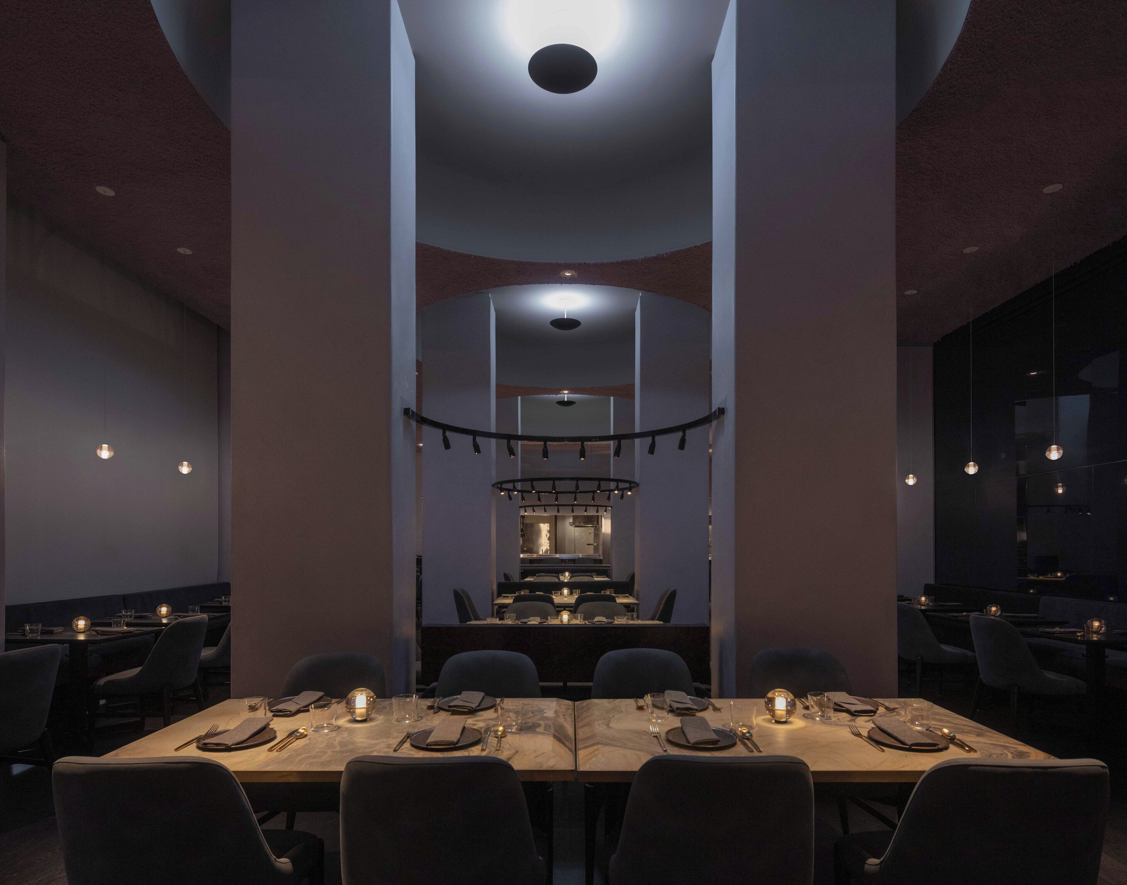 A modern dining area with a long table set for a meal, a kitchen in the background, and pendant lighting.