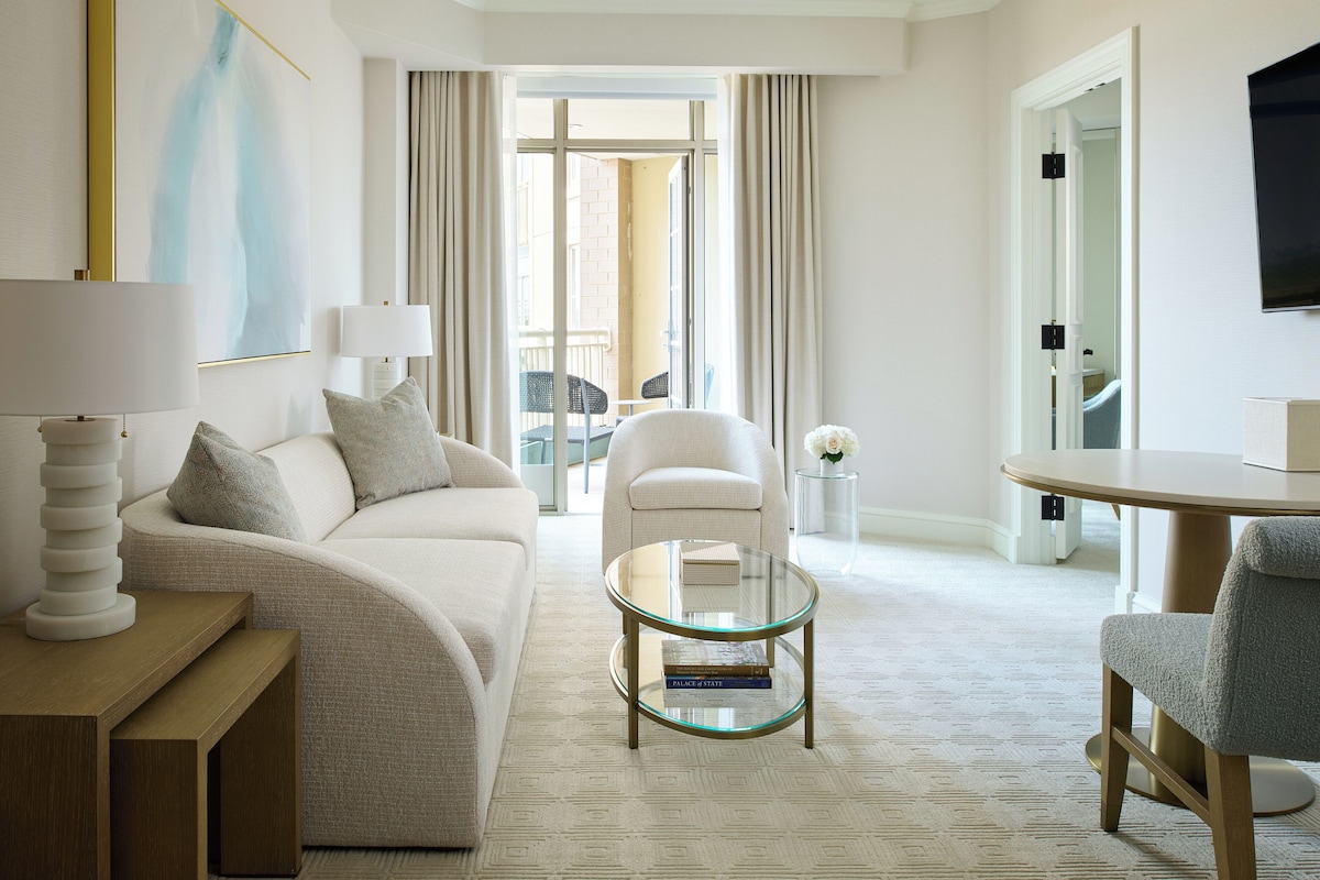 Una habitación de hotel moderna con sofá, mesa de centro de vidrio, mesita auxiliar, una silla y televisor.