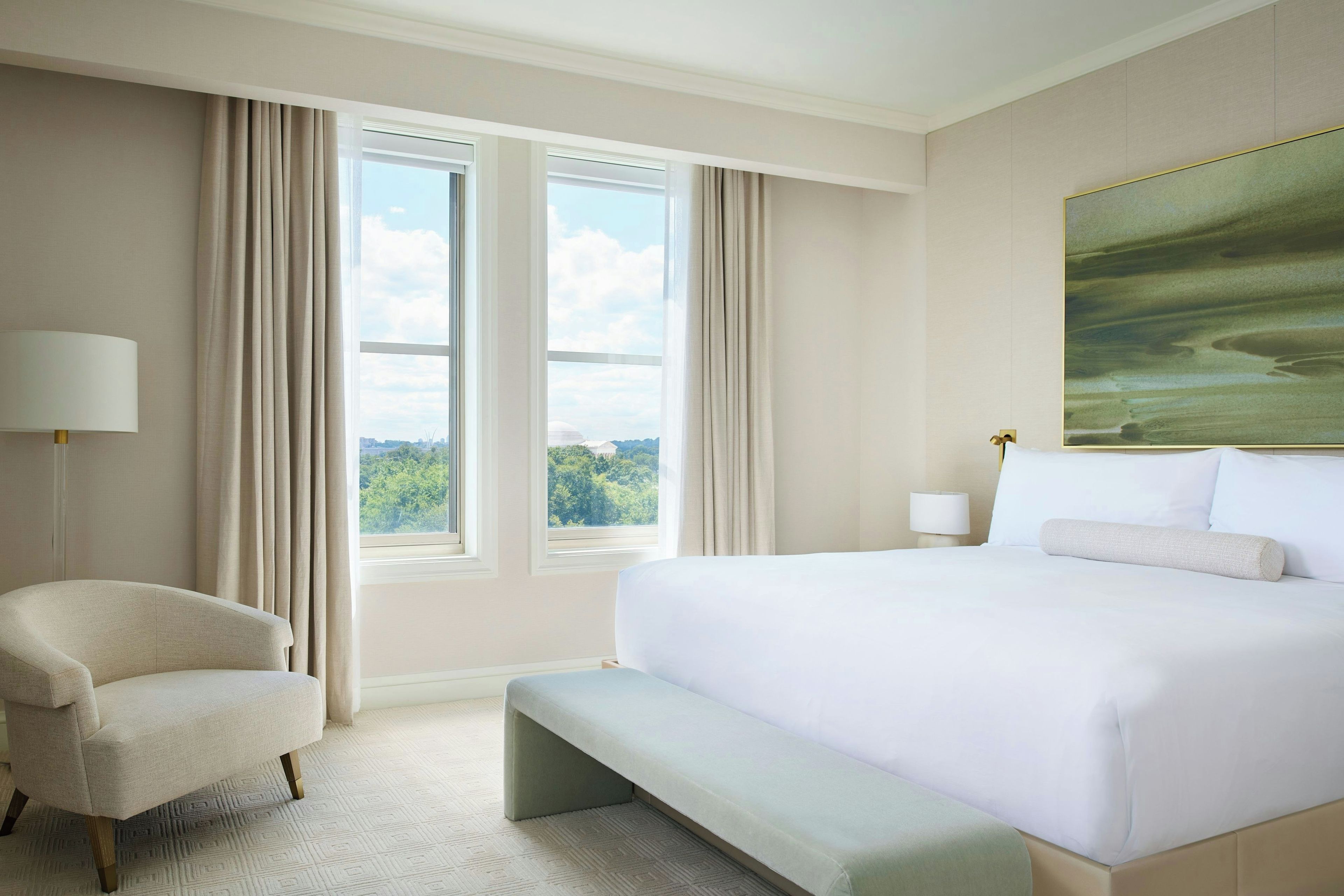 A hotel room with a large bed, a bench, a chair, a nightstand, and a painting on the wall.