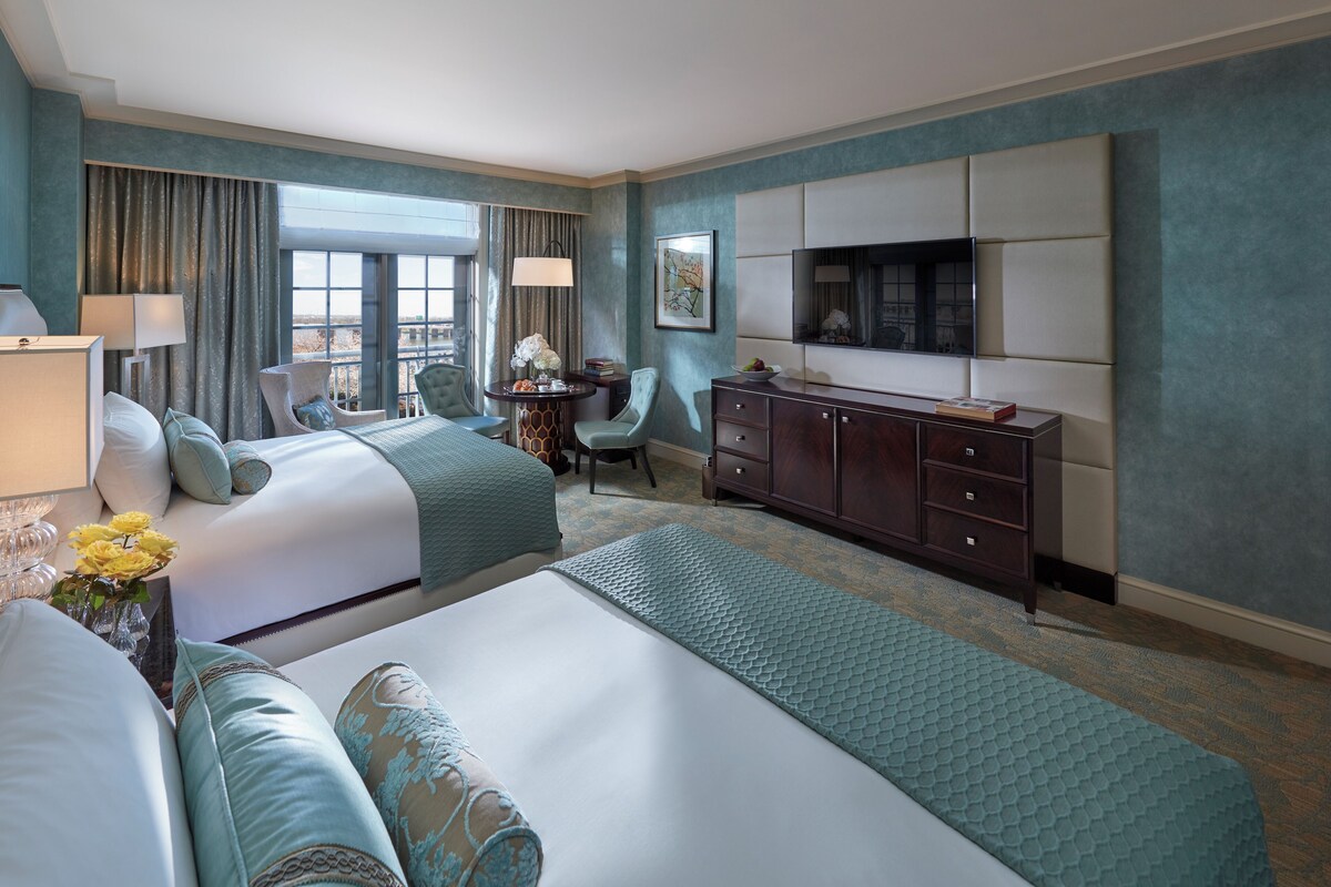 A hotel room with two beds, a desk, a chair, a TV, and a large window.