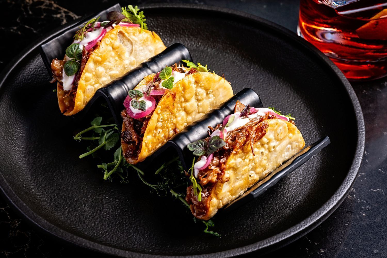 Three tacos on a black plate with a glass of red beverage in the background.