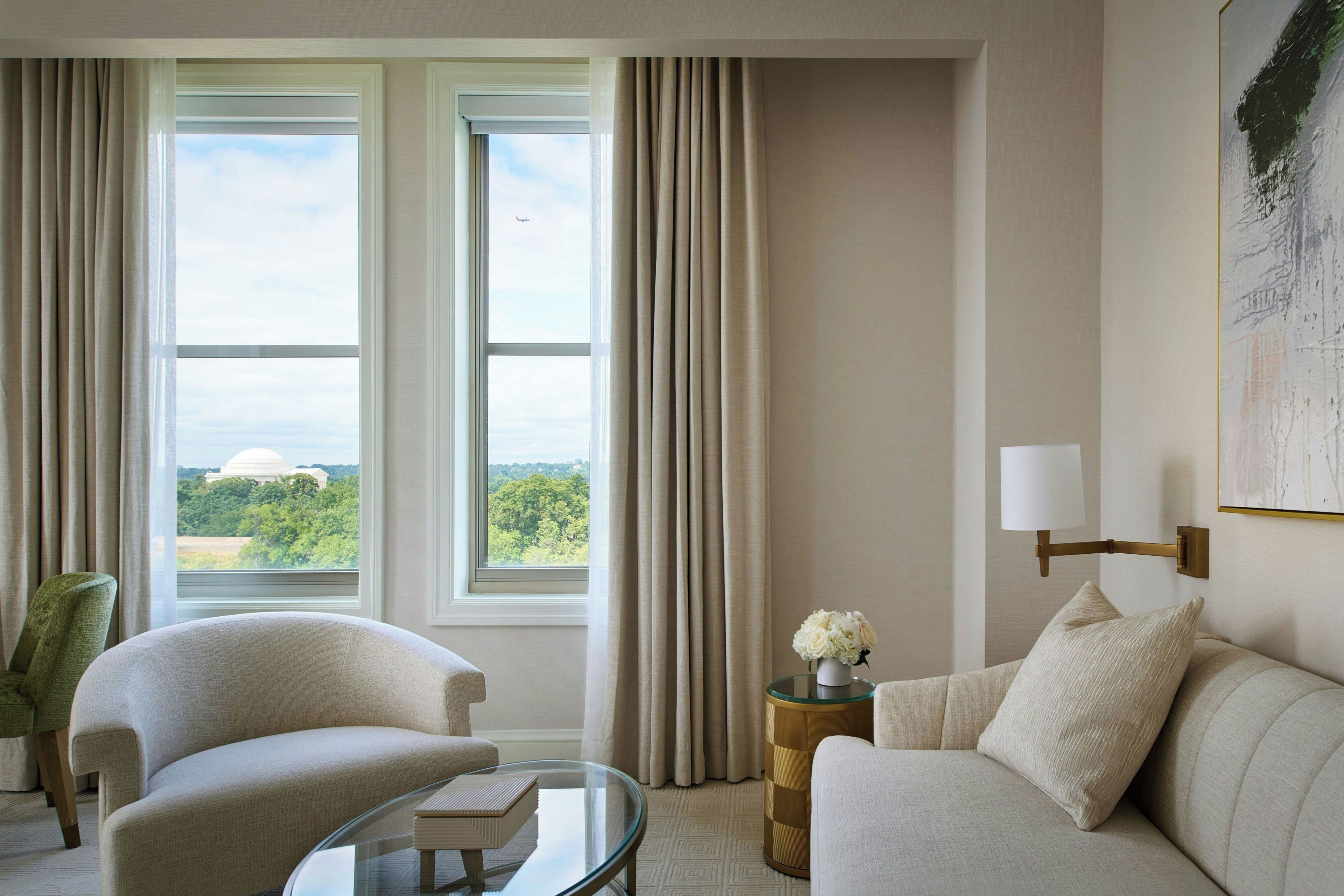 A room with two large windows, a sofa, a chair, a glass coffee table, and a side table with a lamp.