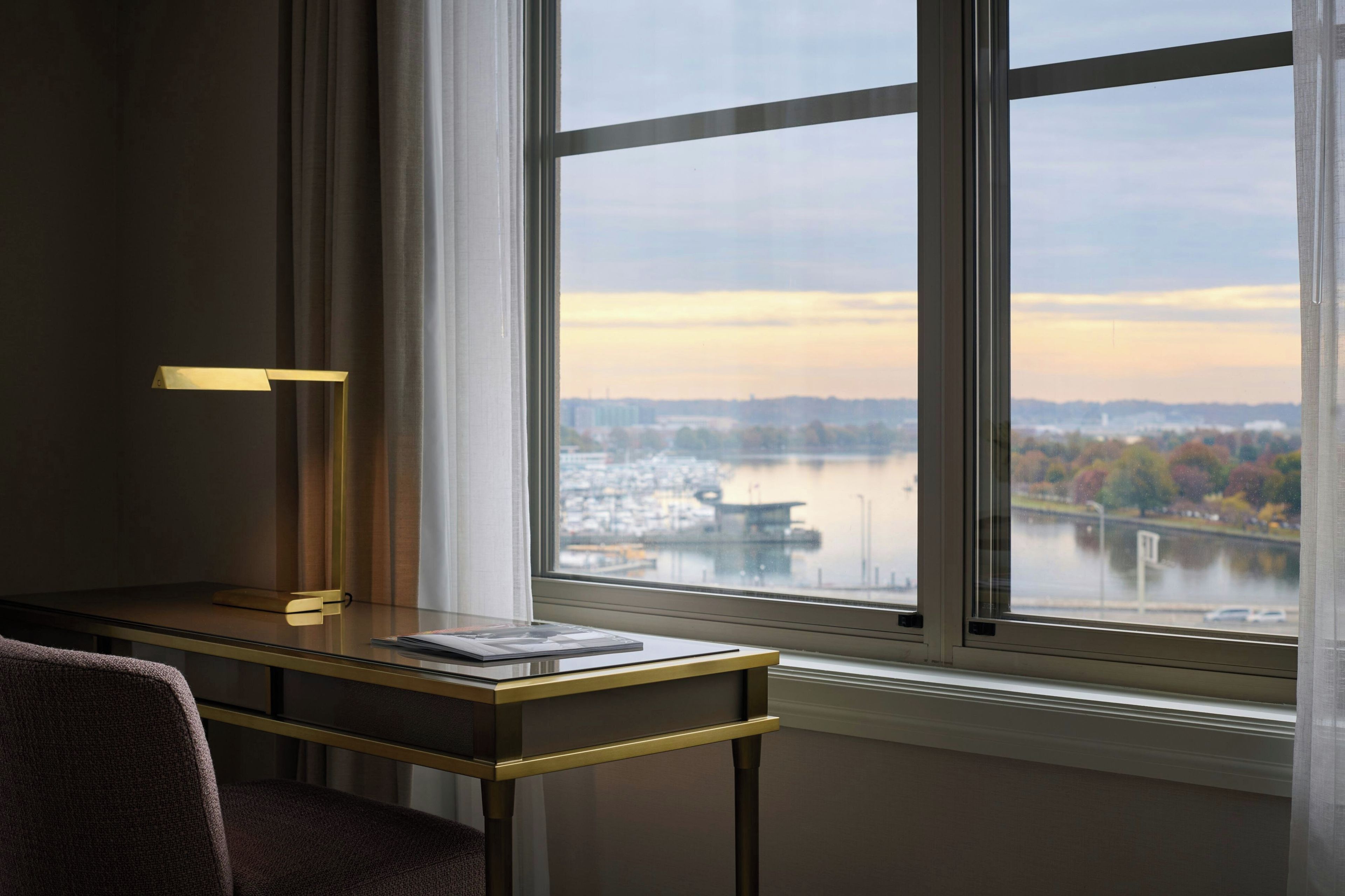 Quarto com escrivaninha, luminária e vista para um rio e paisagem urbana através de uma grande janela.