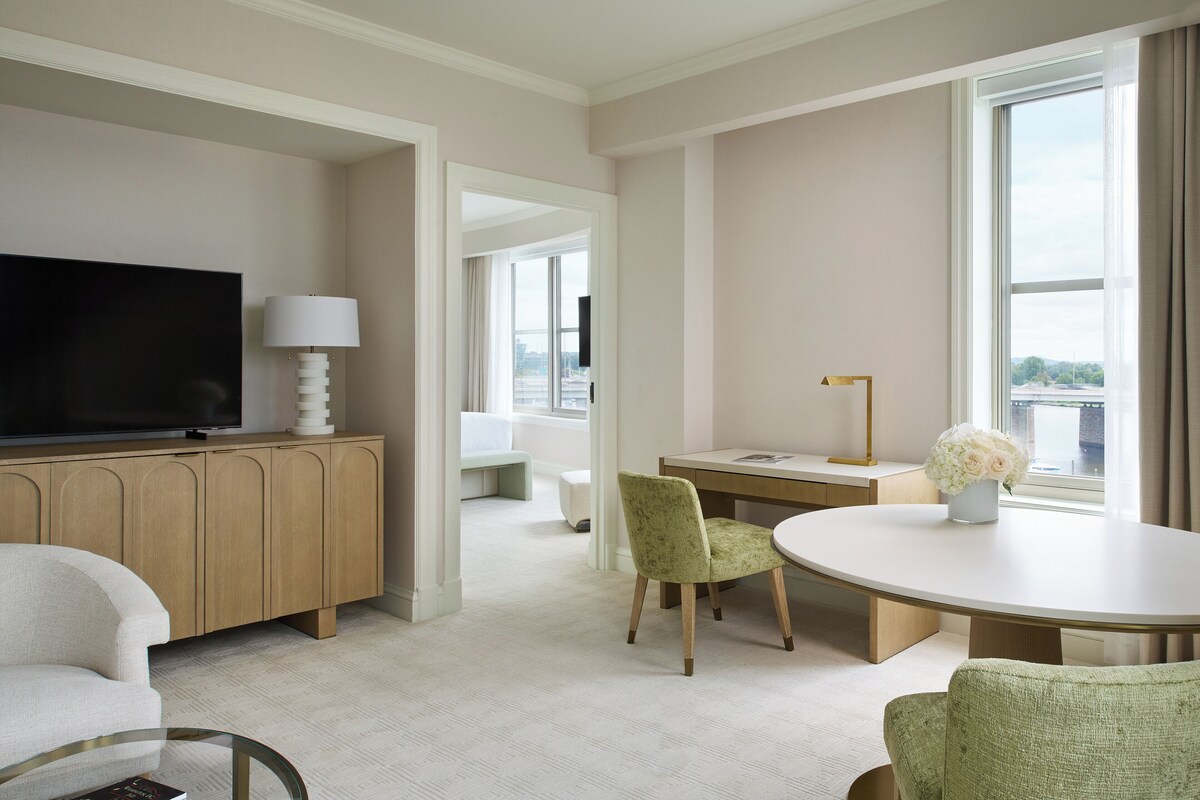 Hotelkamer met flatscreen televisie, houten kast, rond eettafeltje, groene beklede stoel en groot raam met uitzicht.