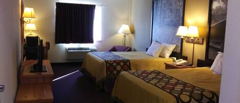 A hotel room with two beds, a desk with a computer, a window with curtains, and a wall-mounted TV.