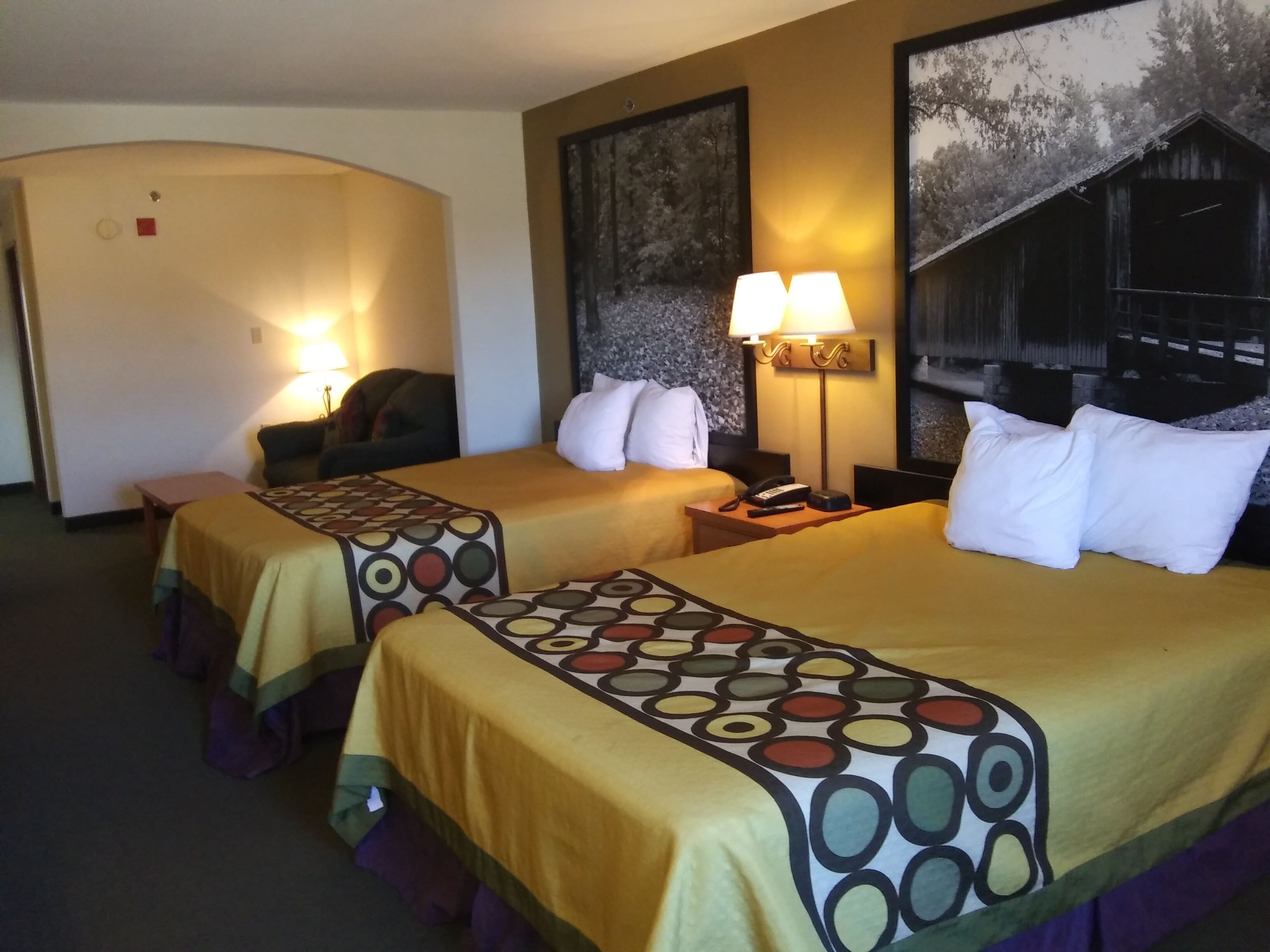 A hotel room with two beds, each with a patterned bedspread and pillows, a desk with a lamp, a chair, and a painting on the wall.