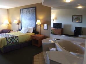 Habitación de hotel con cama, dos lámparas de noche, mesita de noche, televisor, espejo, baño con bañera y un cuadro grande en la pared.