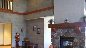 A spacious living room with a stone fireplace, a large window, and a high ceiling.