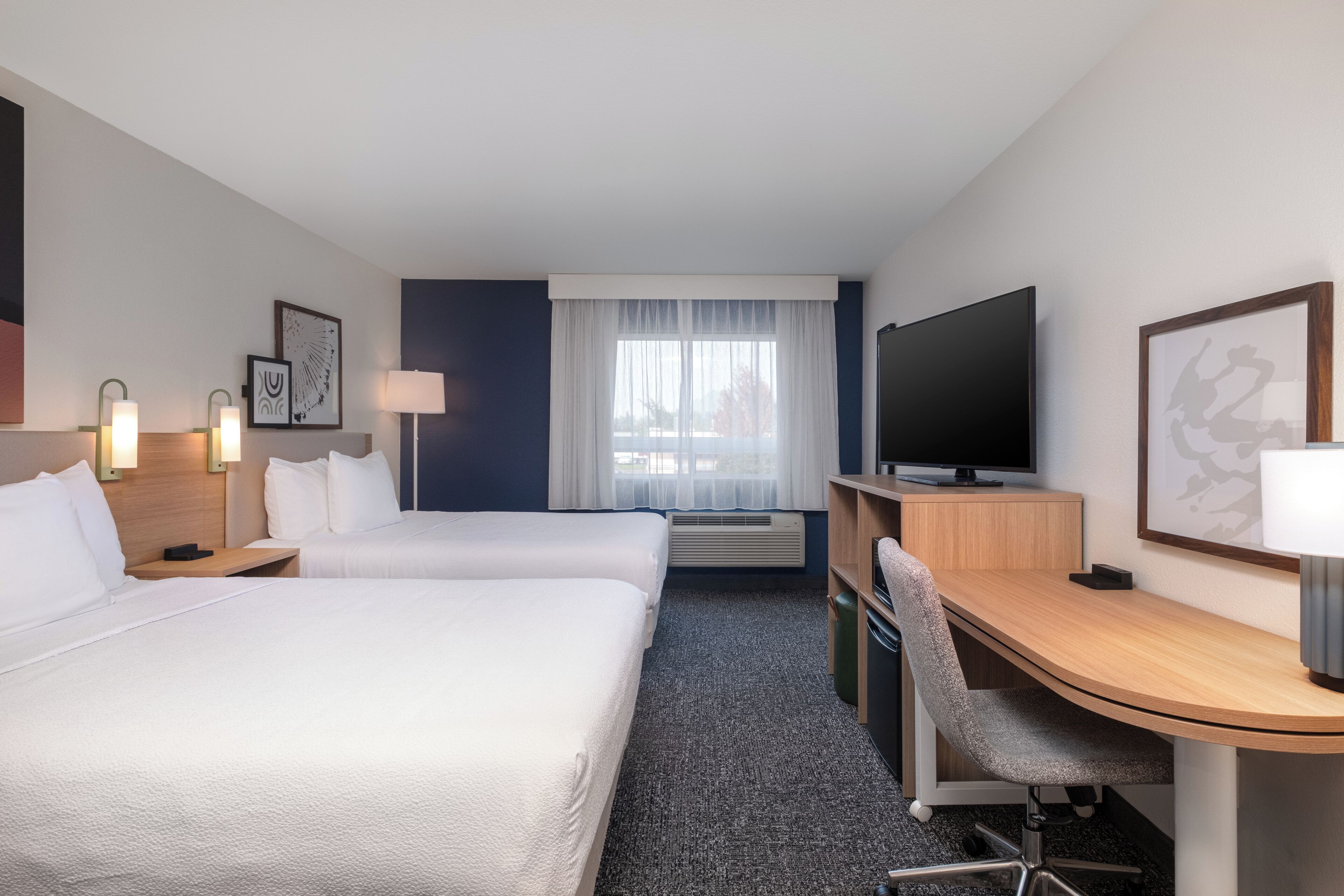 A hotel room with two beds, a desk, a TV, and a window with curtains.