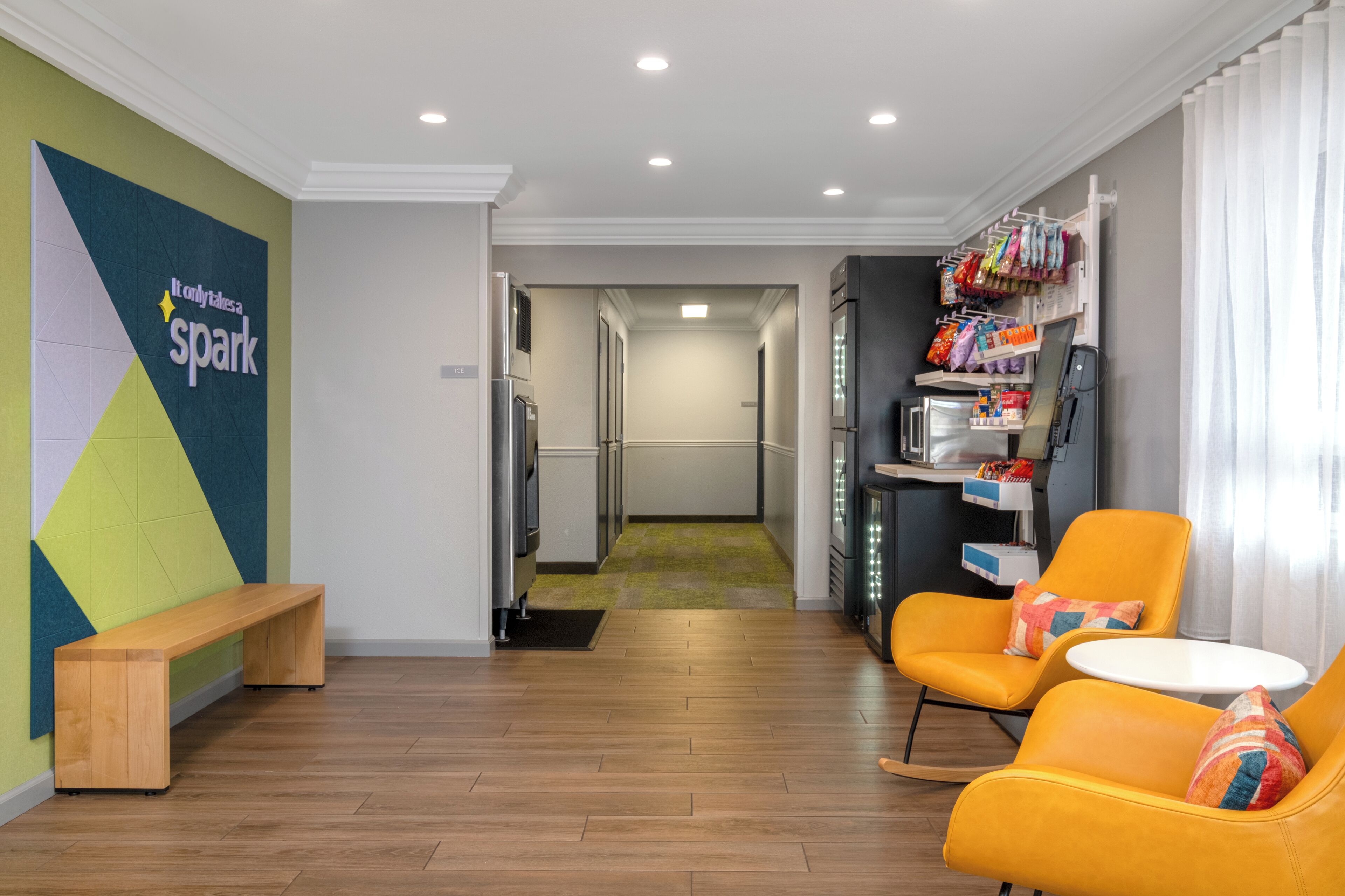 Una habitación de hotel moderna con un banco de madera, un sillón amarillo, una pequeña mesa redonda y una zona de cocina con refrigerador, microondas y un estante con objetos de colores.
