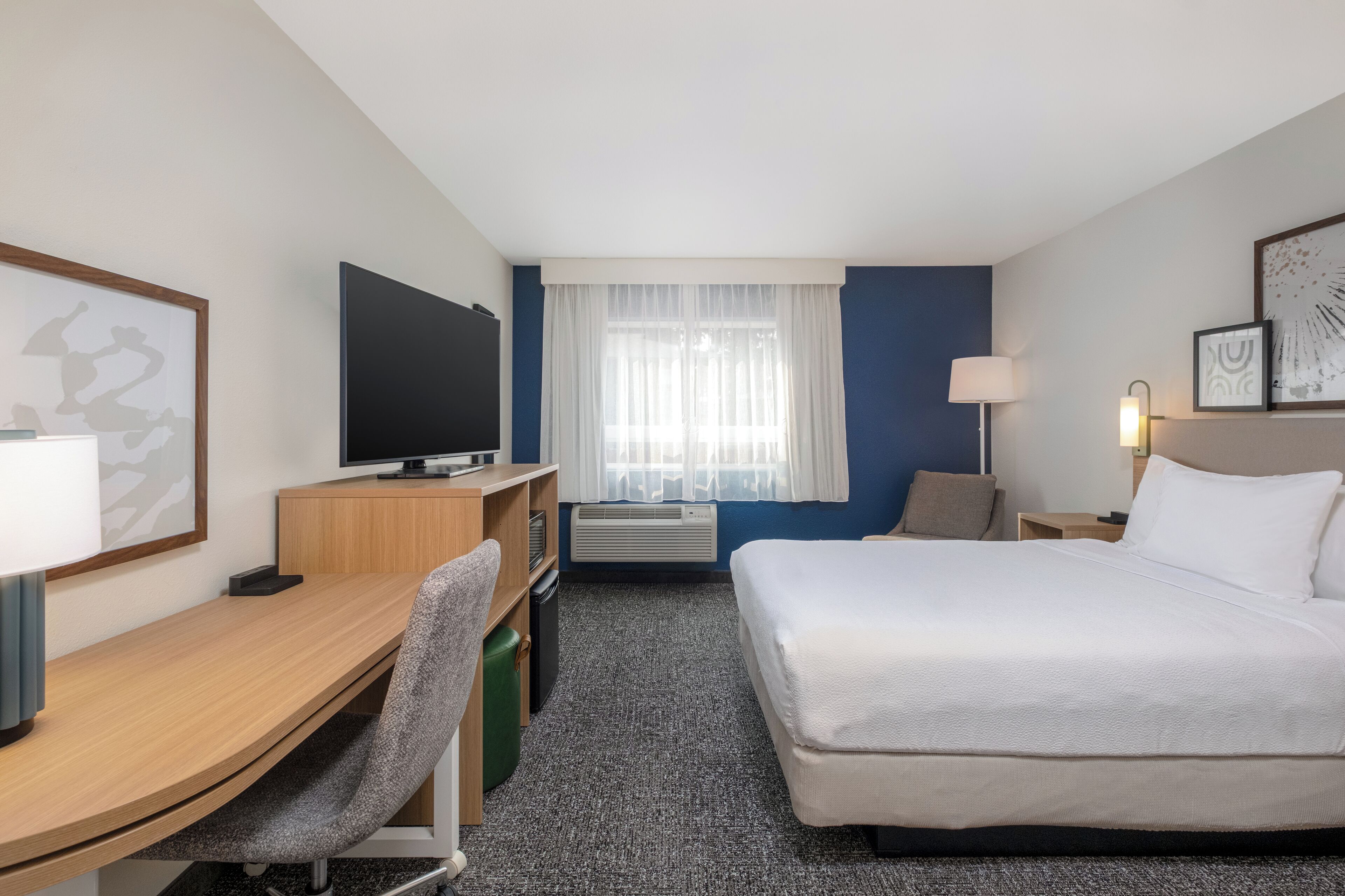 Ein Hotelzimmer mit Bett, Schreibtisch, Stuhl, Fernseher und einem Fenster mit Vorhängen.