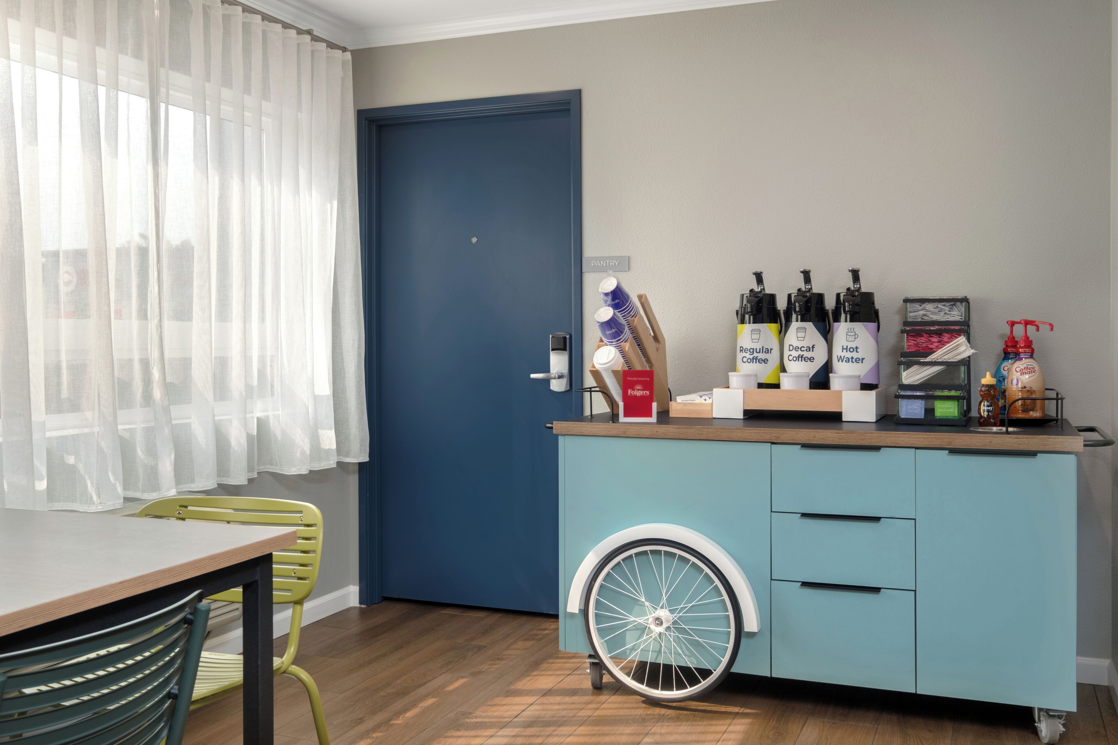A room with a blue door, a kitchenette with a wheelbarrow, a dining table, and chairs.