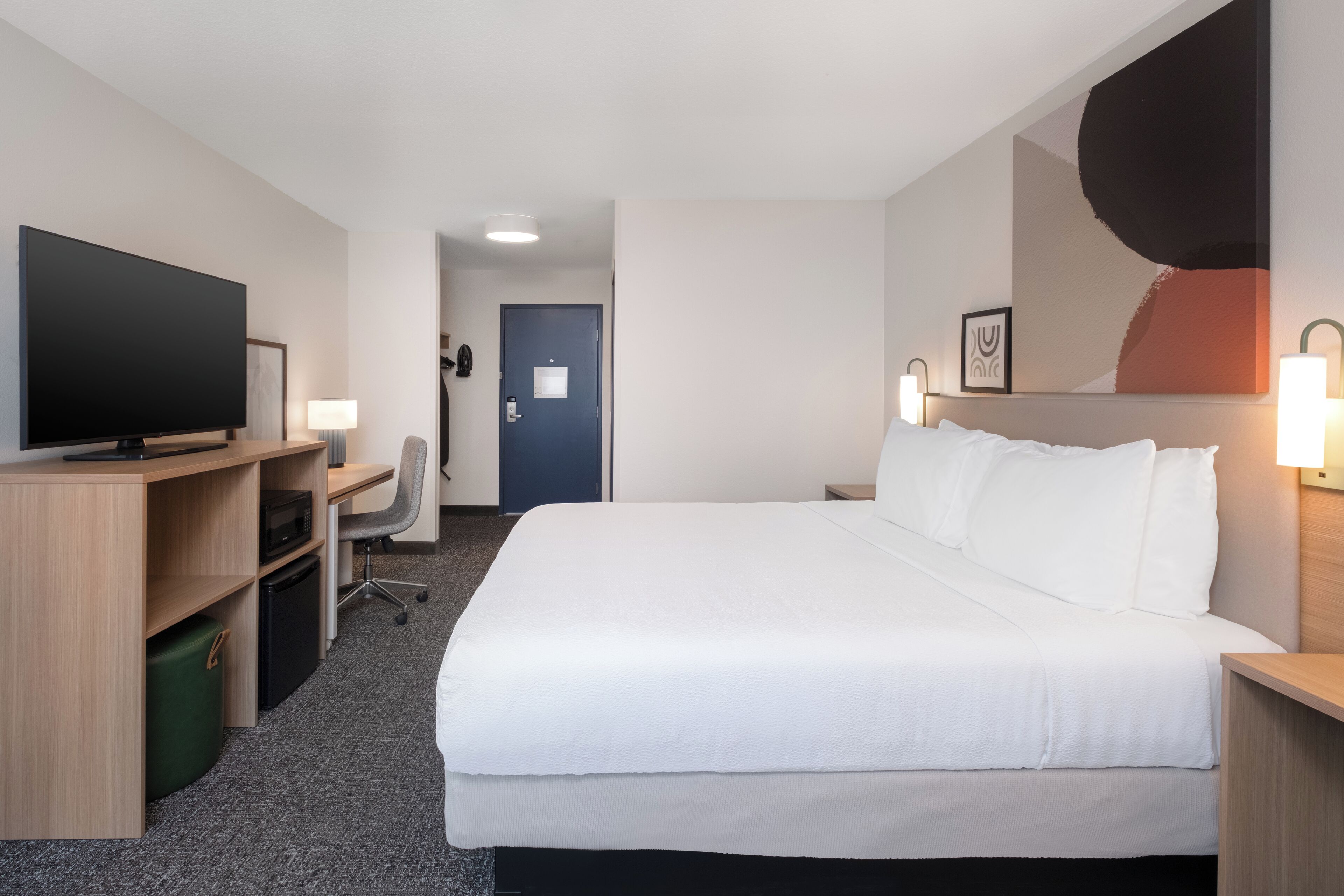 A hotel room with a large bed, a desk with a flat-screen TV, a chair, and a small desk with a lamp.