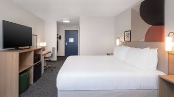 A hotel room with a large bed, a desk with a flat-screen TV, a chair, and a small desk with a lamp.