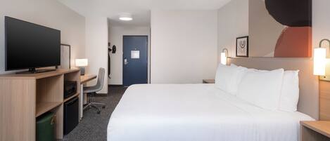 A hotel room with a large bed, a desk with a flat-screen TV, a chair, and a small desk with a lamp.