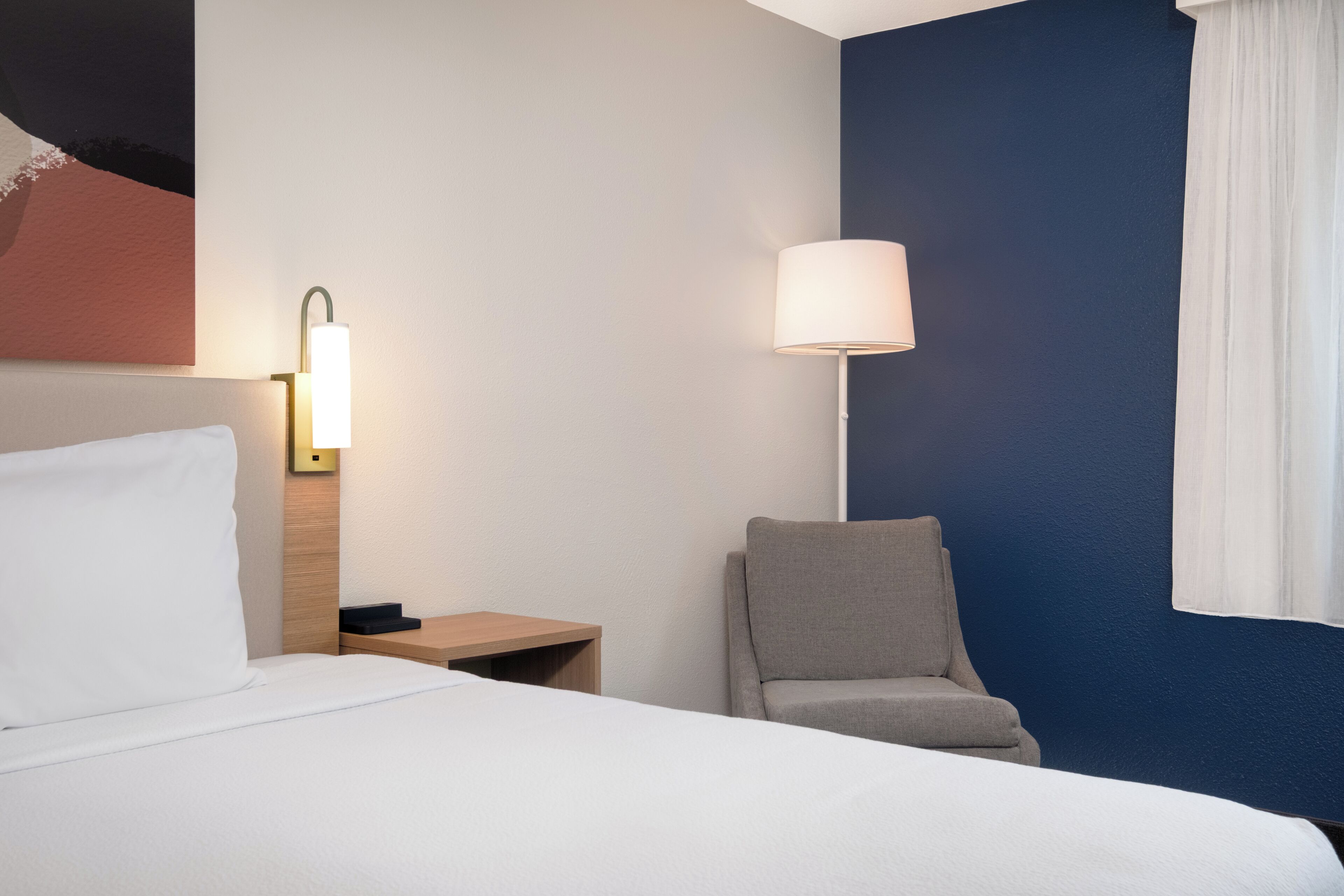 A hotel room with a bed, a chair, a nightstand, and a wall-mounted lamp.