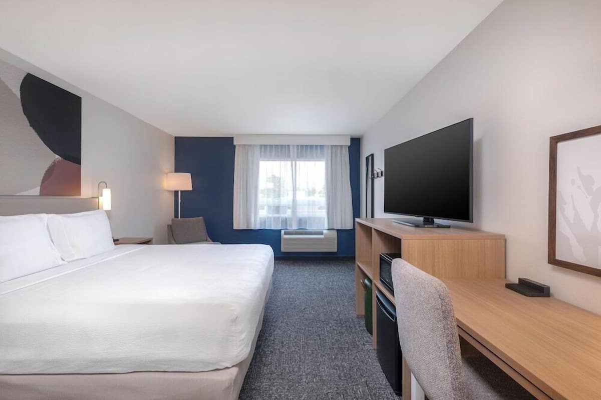 Una habitación de hotel con cama, escritorio, televisor y ventana con cortinas.