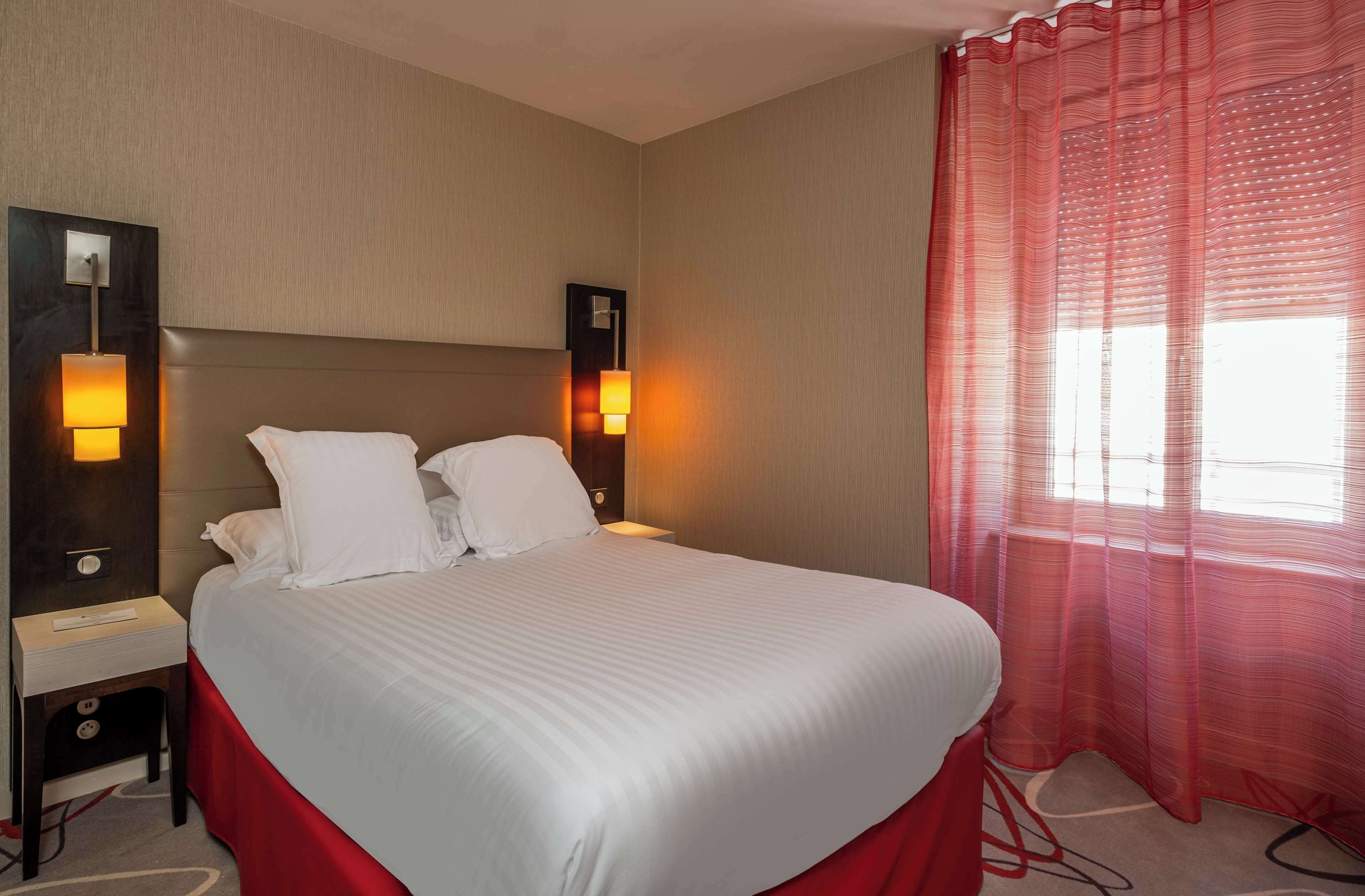 A hotel room with a large bed, two bedside lamps, a nightstand, and a window with sheer curtains.