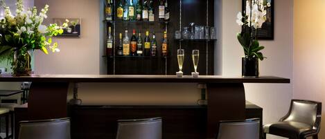 Una zona de bar con mostrador oscuro, tres taburetes, una variedad de botellas de licor y dos copas de champán.